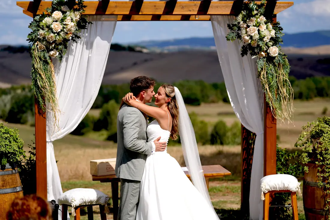 Bride and groom kissing under the flower arch with the Tuscan hills behind